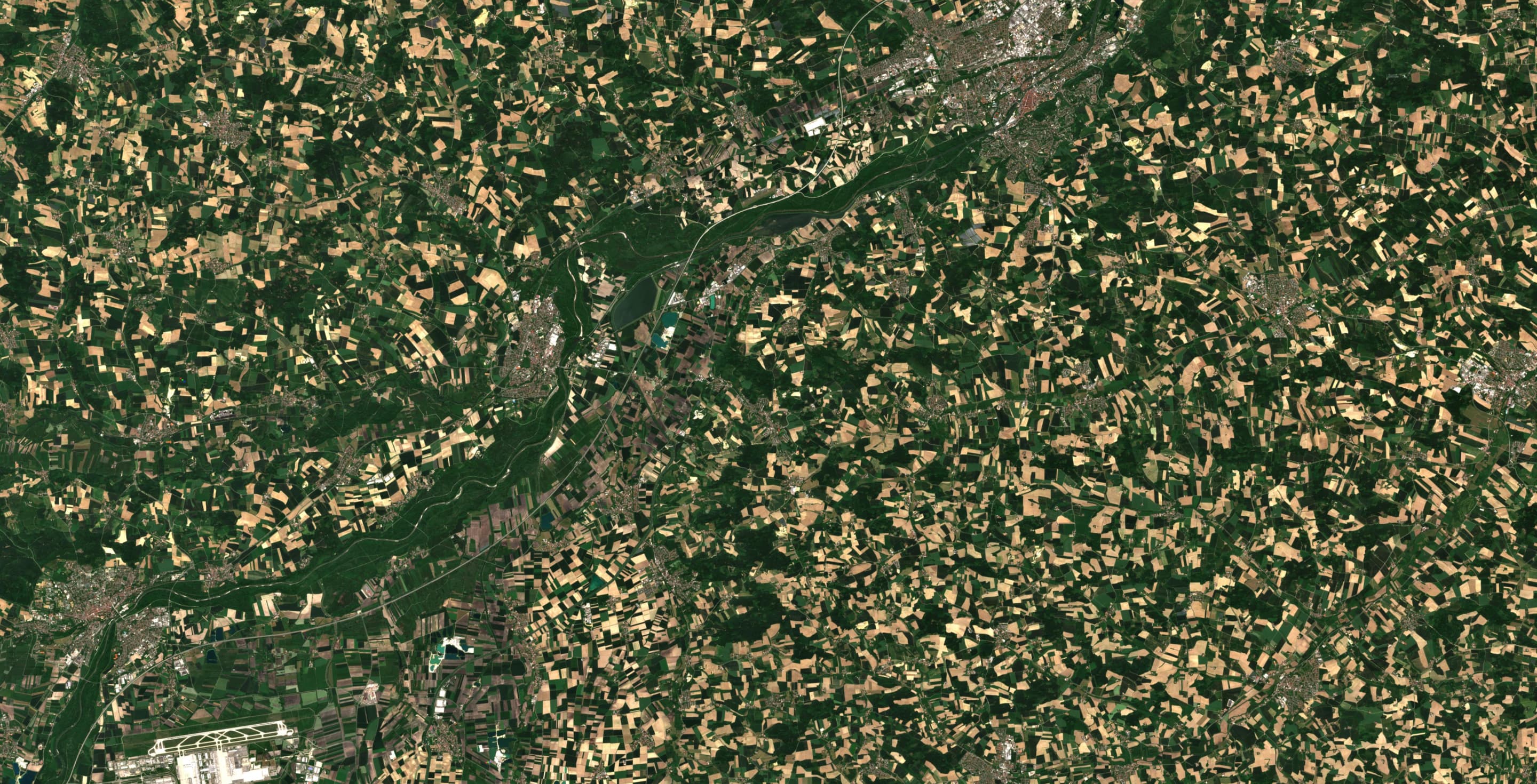 A satellite image showing a river flowing through rural agricultural fields, with patches of green and yellow land, roads, and nearby urban areas.