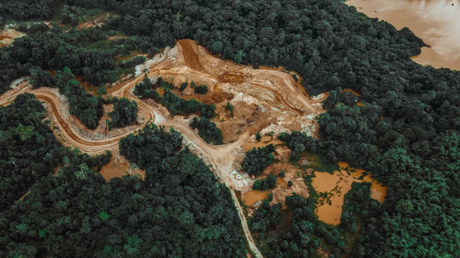 Aerial view of deforestation in the Amazon rainforest, showing a large cleared area with exposed soil, winding dirt roads, and surrounding dense forest.