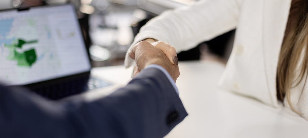 Close-up of two professionals shaking hands over a desk, with a laptop displaying a map in the background