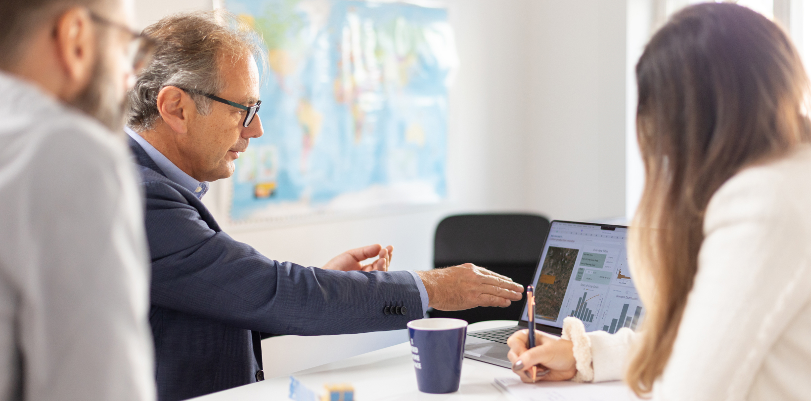 A professional team reviewing data charts on a laptop during a business meeting, with a world map in the background