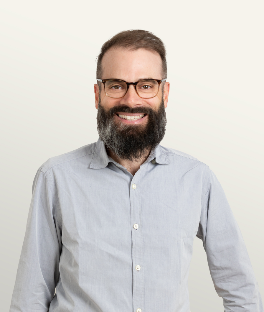 A bearded man with glasses wearing a light gray shirt, smiling confidently against a neutral background.