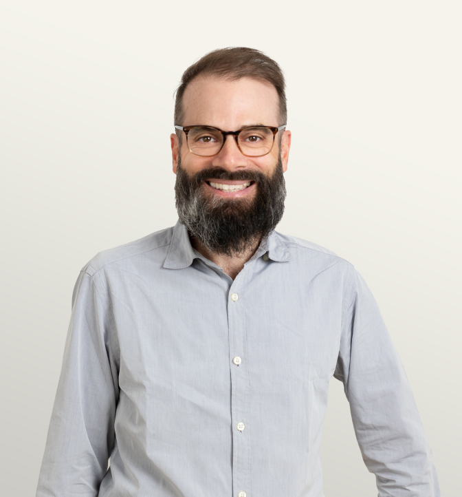 Portrait of a confident man with a full beard, glasses, and a light gray button-up shirt, smiling against a neutral background