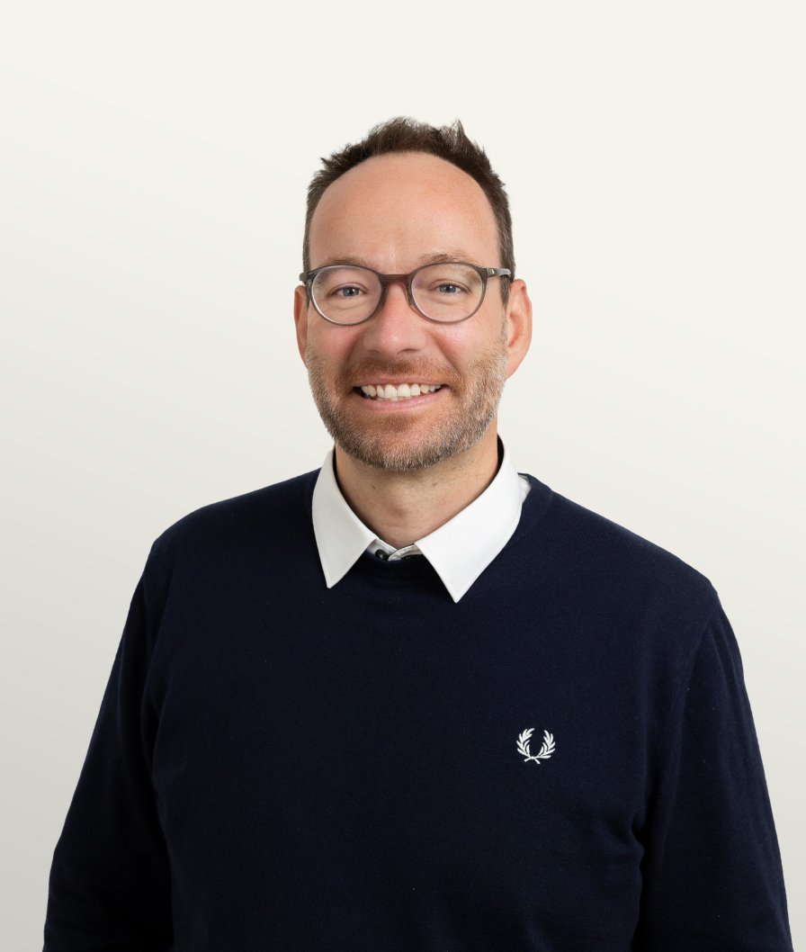 Portrait of a smiling business professional wearing glasses, a navy sweater, and a white collared shirt against a neutral background.