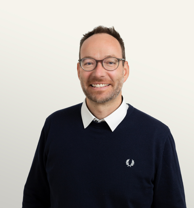 Portrait of a confident man with short brown hair, glasses, and a dark sweater with a white collar, smiling against a neutral background