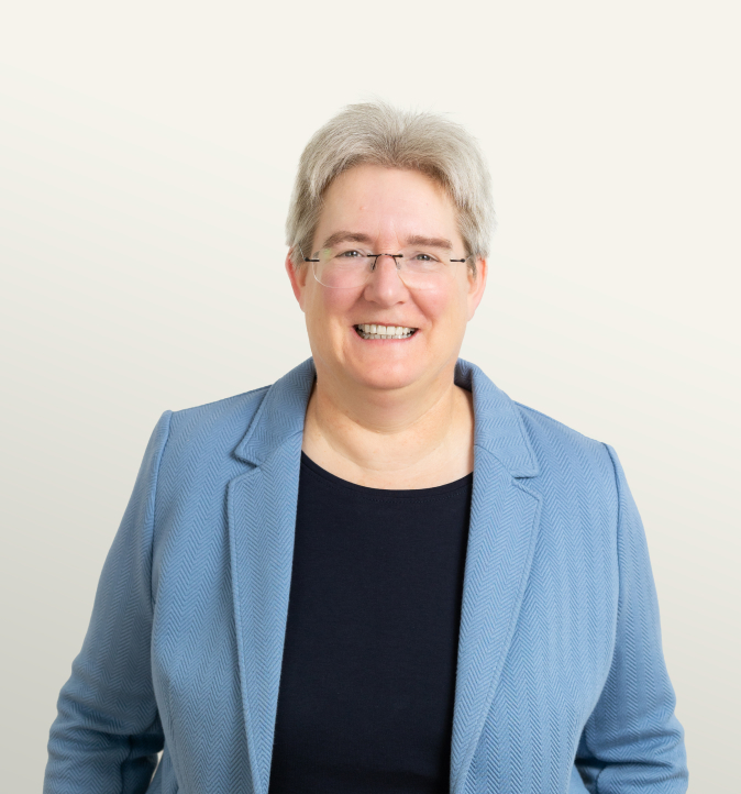 Portrait of a confident woman with short gray hair, wearing glasses and a blue blazer, smiling against a neutral background