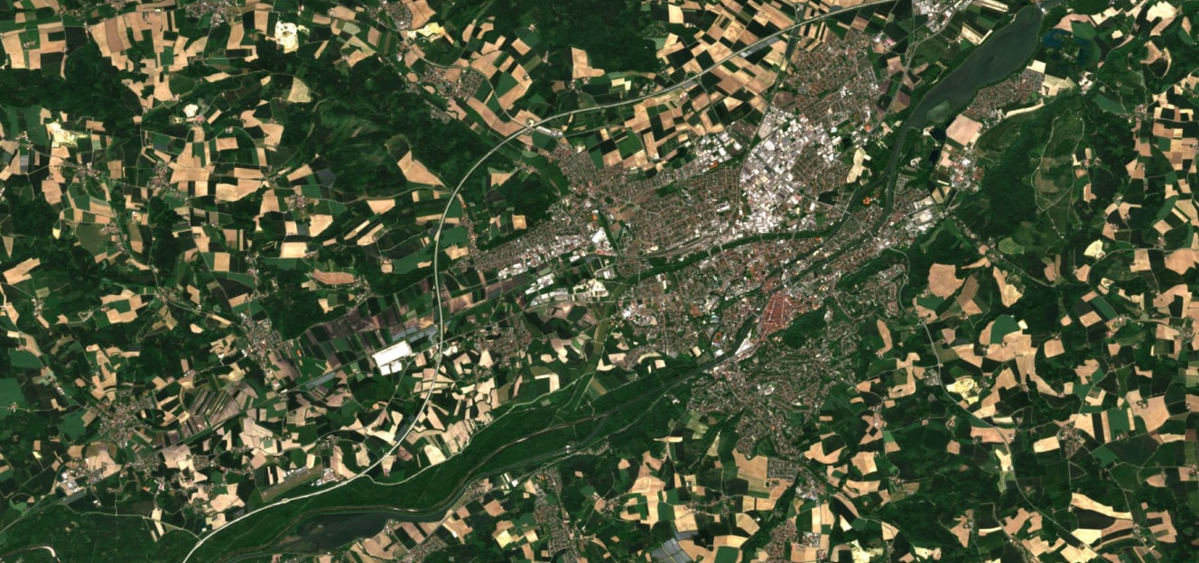 High-resolution satellite image showing a mix of farmland, forests, and urban settlements with a river running through the landscape