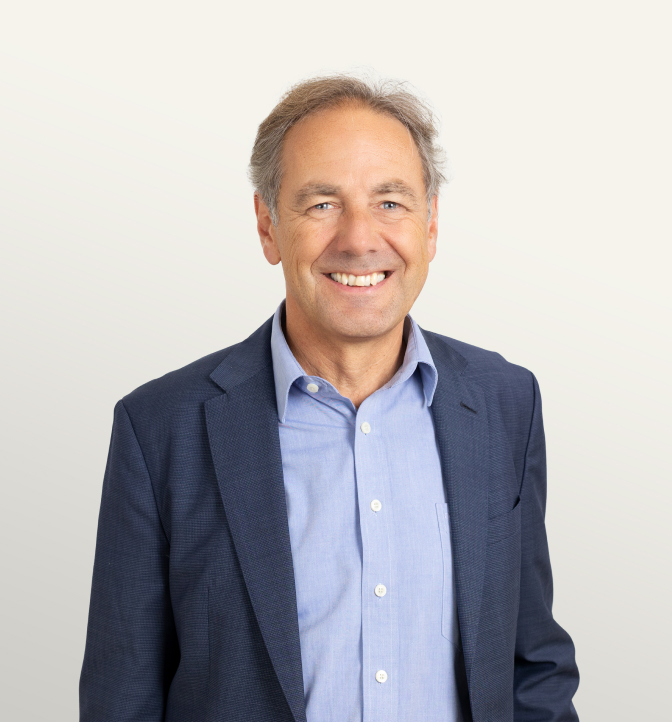 Portrait of a confident man with short gray hair, wearing a navy blue blazer and a light blue shirt, smiling against a neutral background