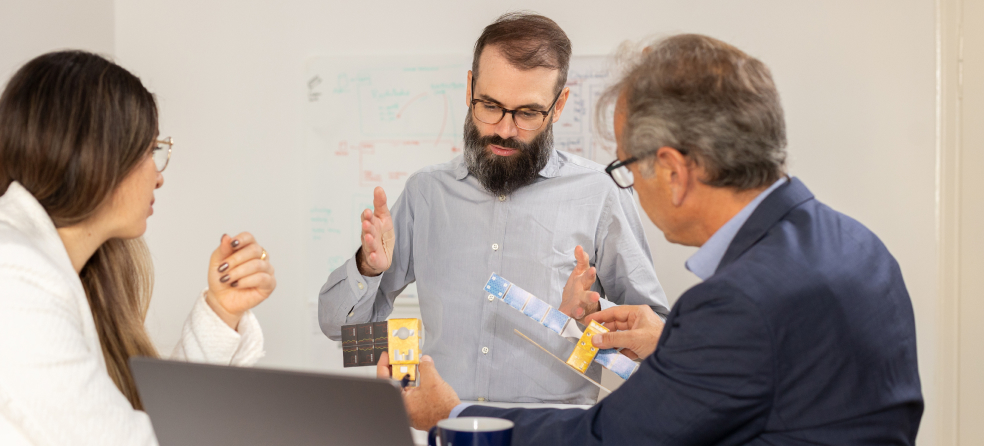 Three professionals in a meeting analyzing a satellite model with technical sketches in the background