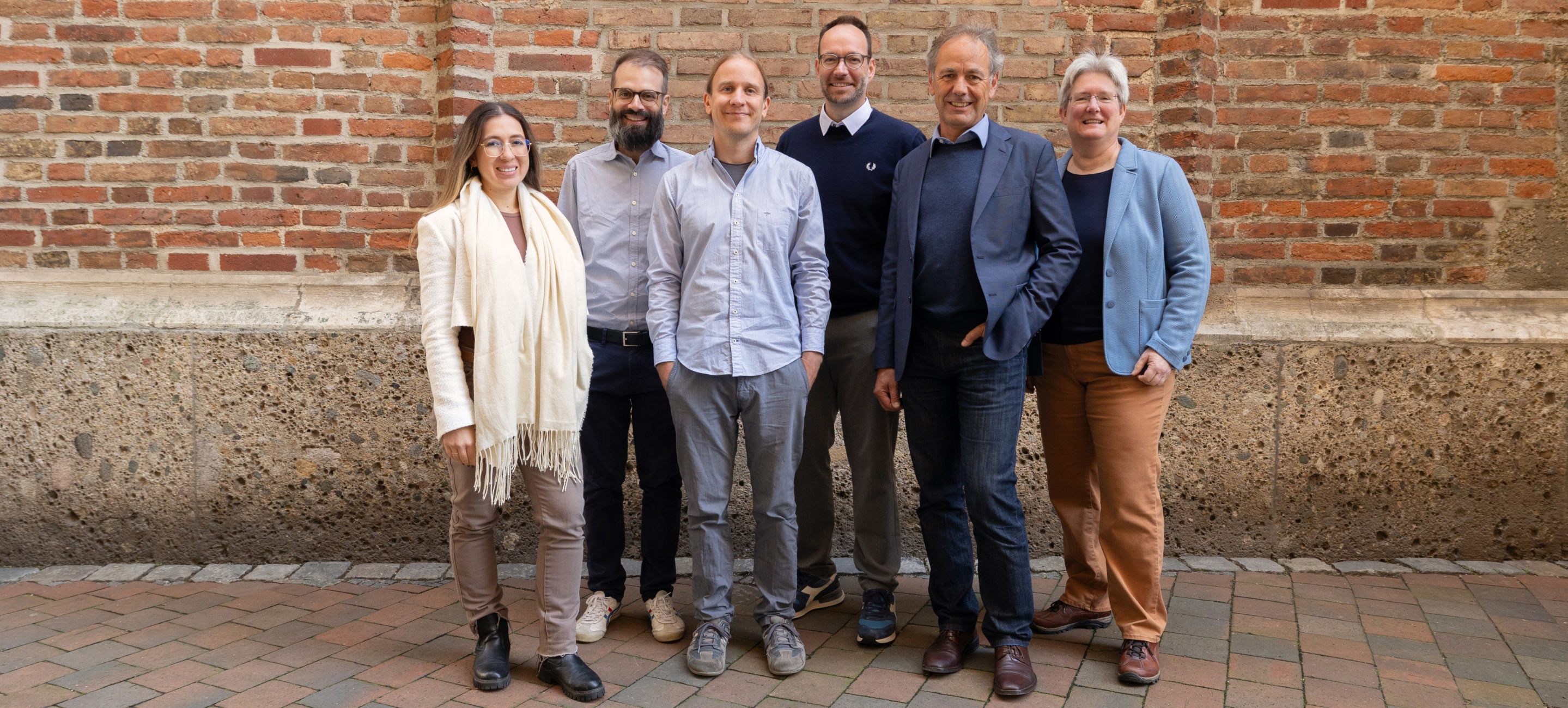 Six team members standing together outdoors, smiling in front of a rustic brick wall